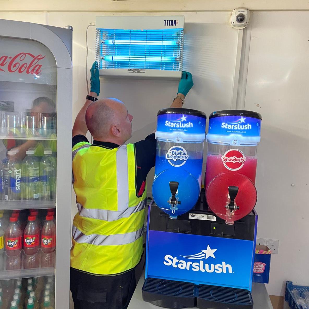 A Don Valley pest controller in a high-vis vest adjusting an electric fly trap in a food business, stood between a vending machine full of drinks and a slushy machine.